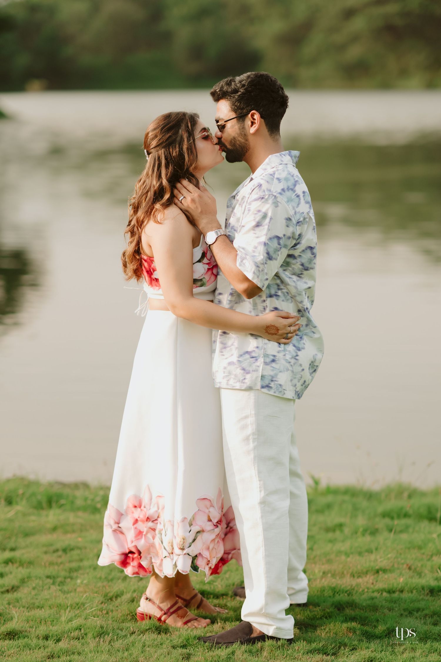 A romantic kiss between a couple in matching white outfits by a serene lakeside setting.