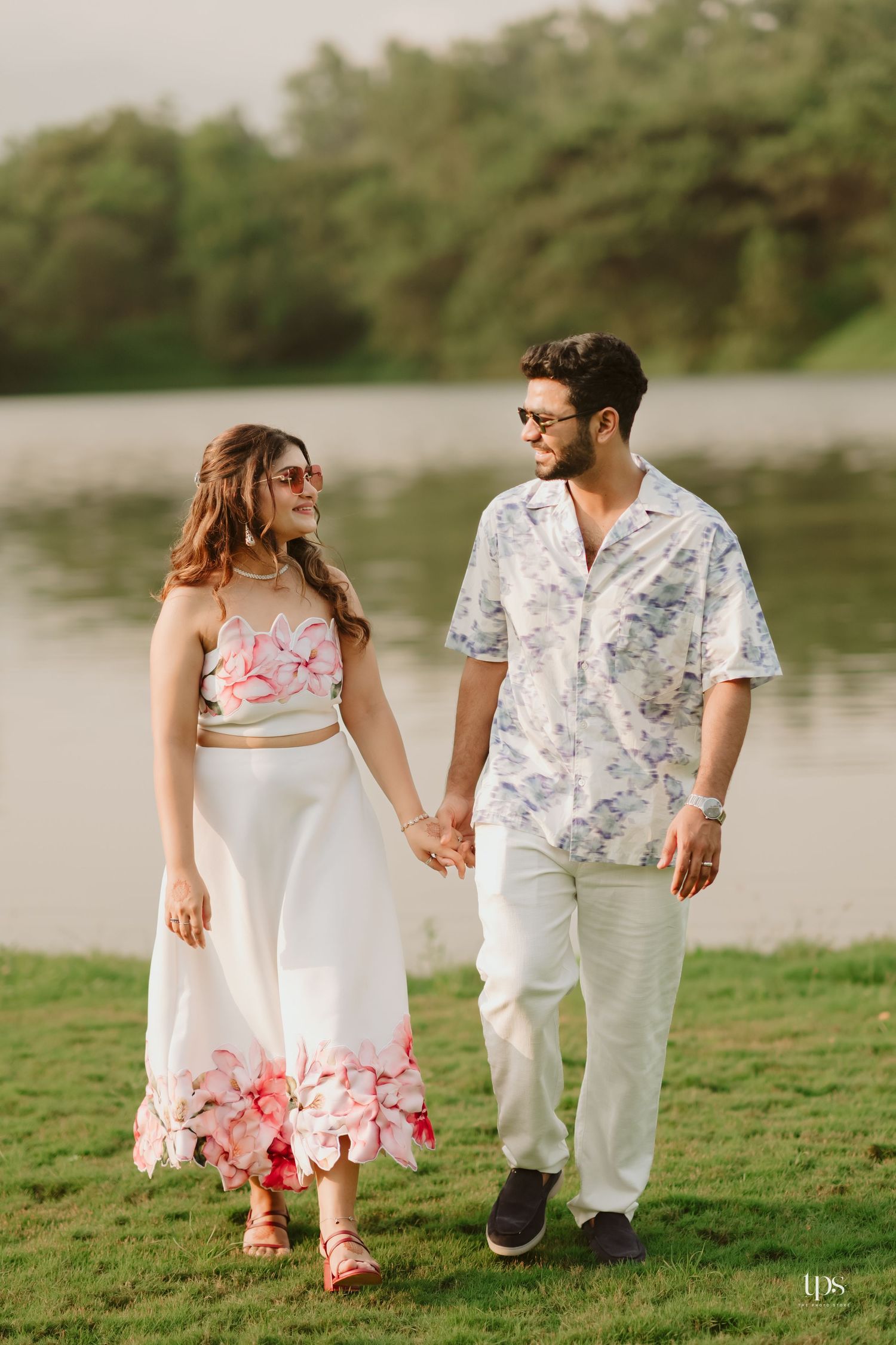 A couple takes a romantic stroll holding hands, wearing coordinated white and floral outfits by a lake.