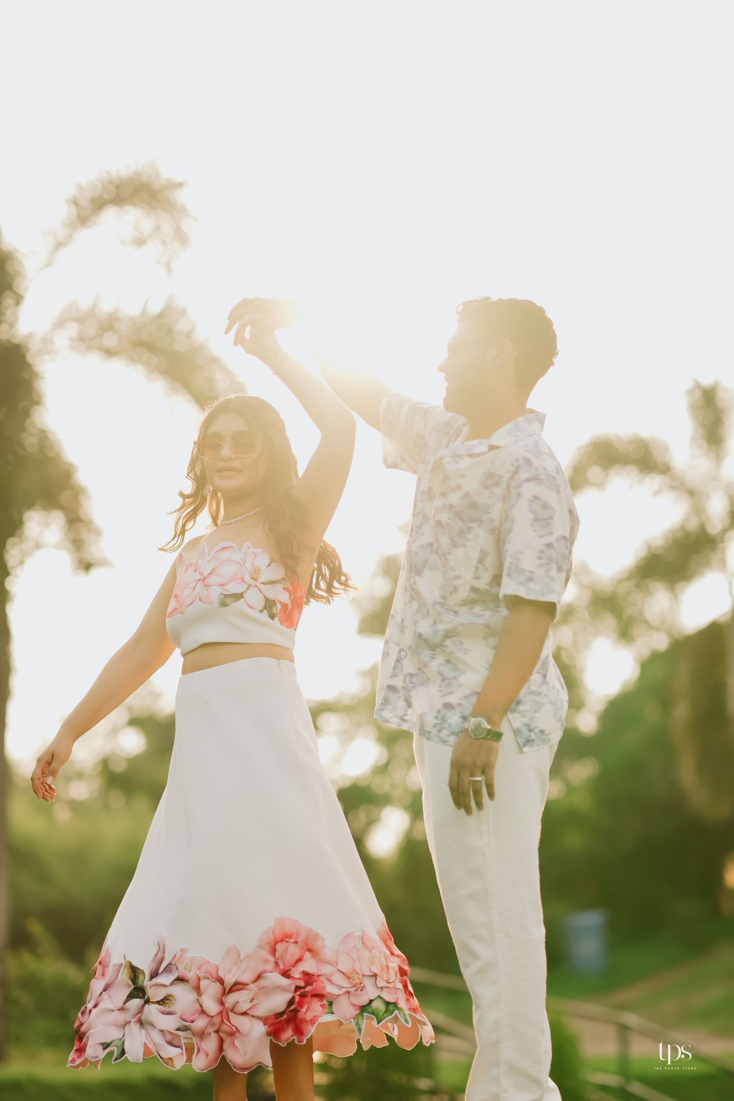 A couple dances together in a sunlit outdoor setting, wearing matching white and floral attire.