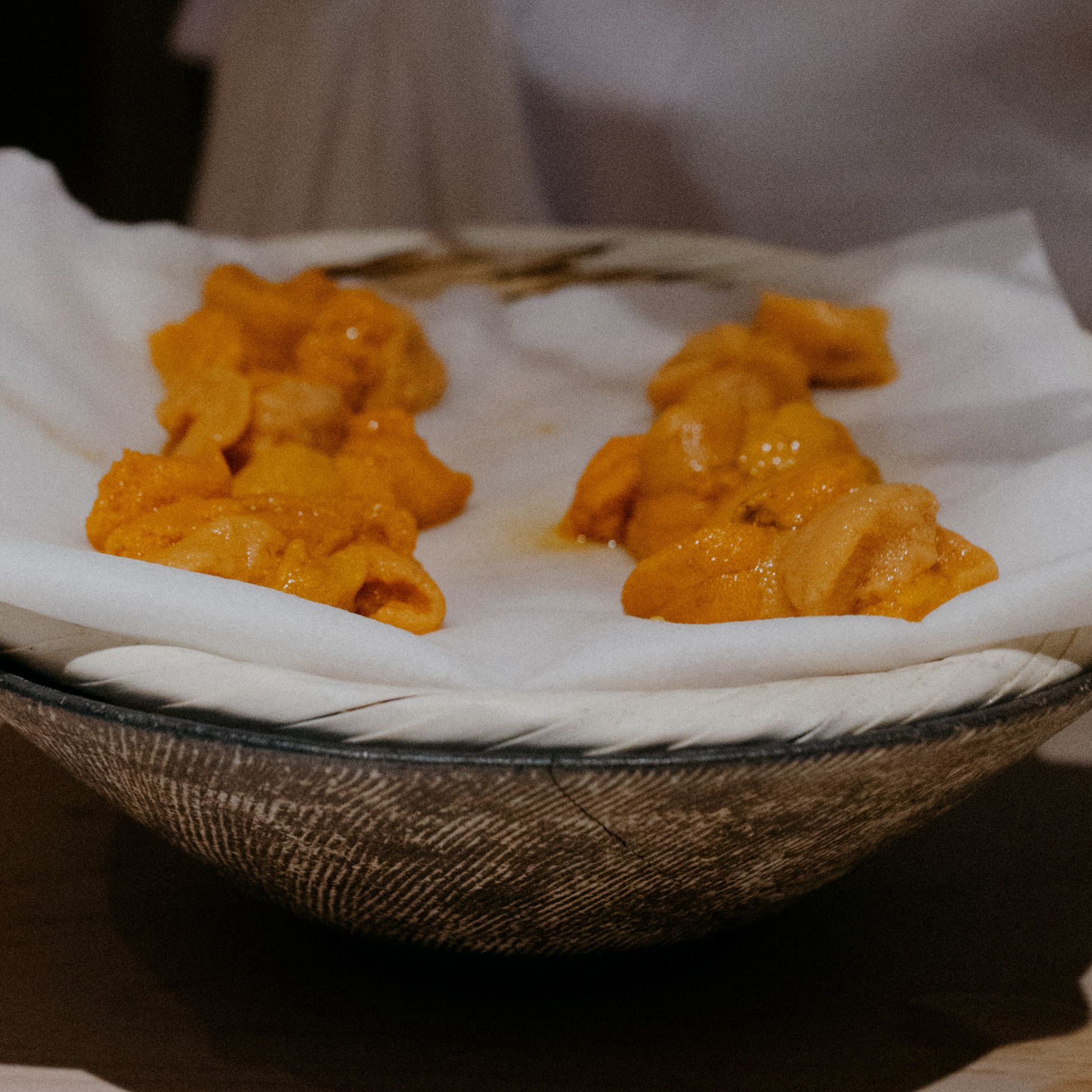 Orange uni sea urchin pieces arranged on white paper in a rustic bowl.
