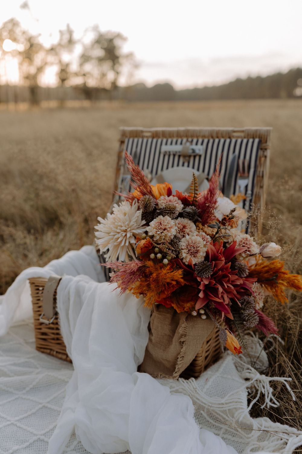 Rustic wicker picnic basket with autumn floral arrangement and white fabric in a golden field at sunset.