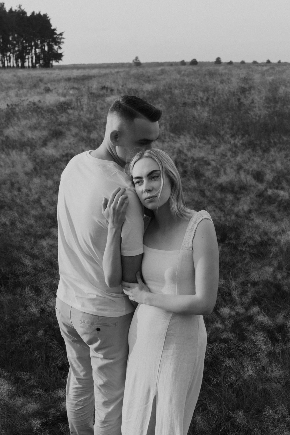 Romantic couple in white outfits standing together in a field at sunset.