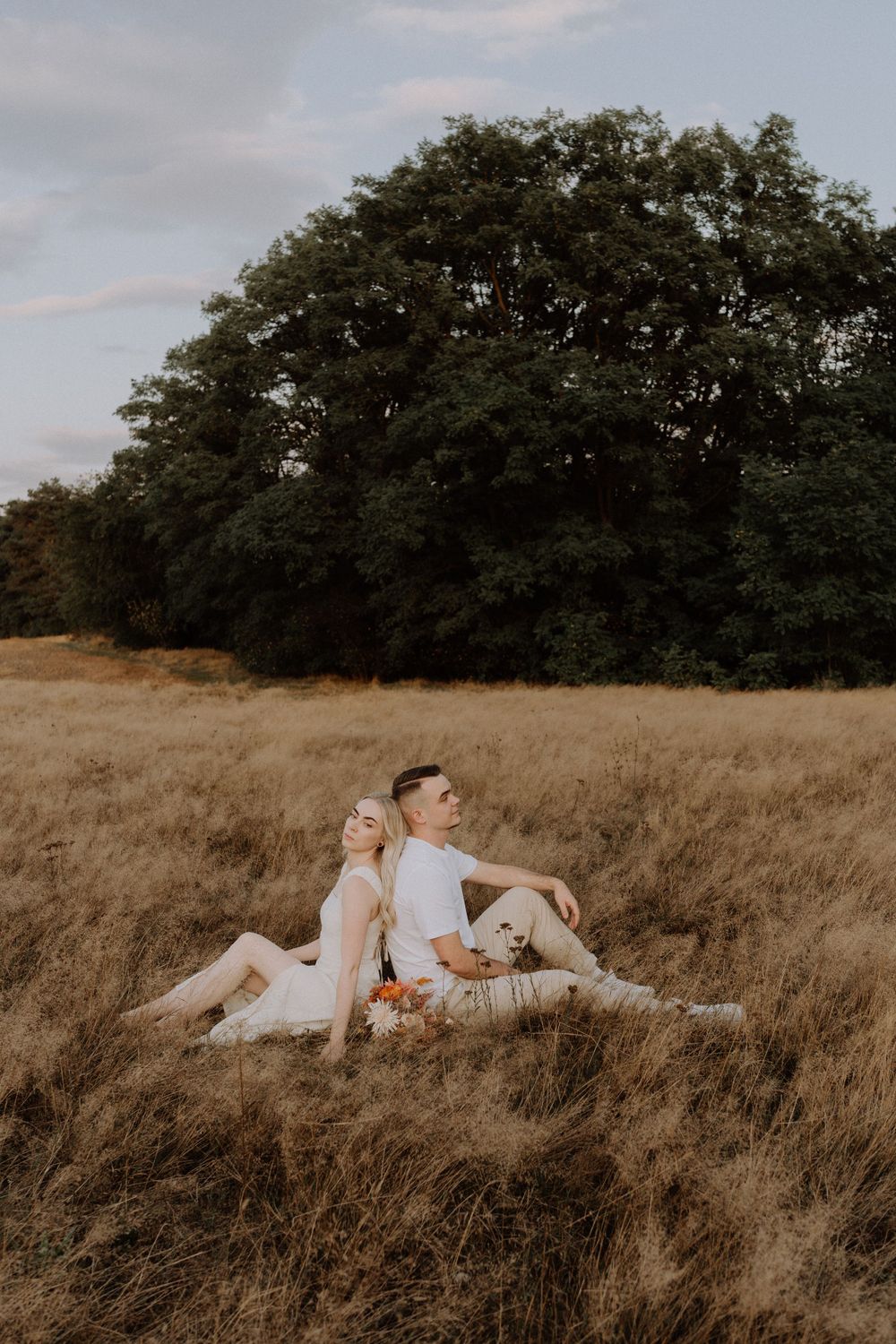 Couple sitting back-to-back in a sunlit meadow with trees in background.