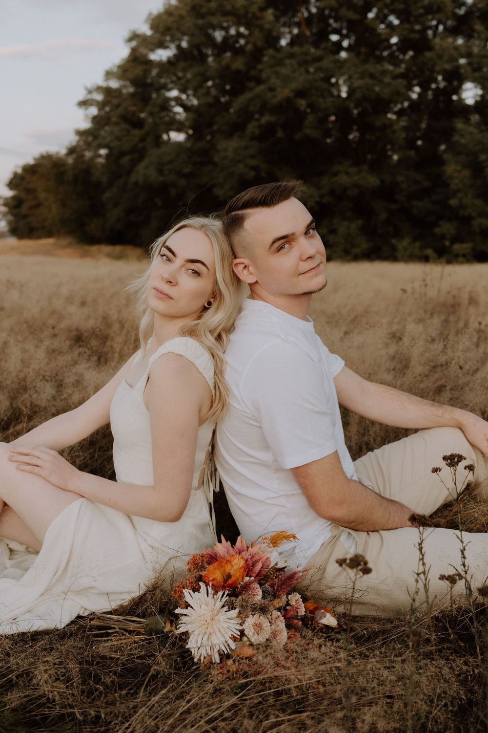 Couple in white outfits sitting back-to-back with autumn bouquet in field.
