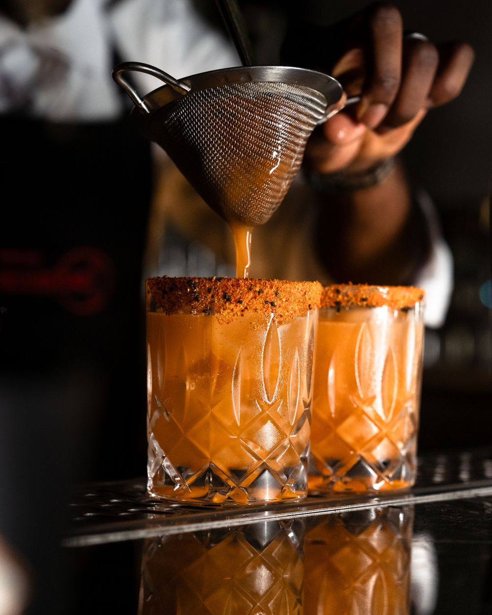 Craft cocktail being strained through fine mesh strainer into crystal cut glass with orange liquid. Mtlshutter photography