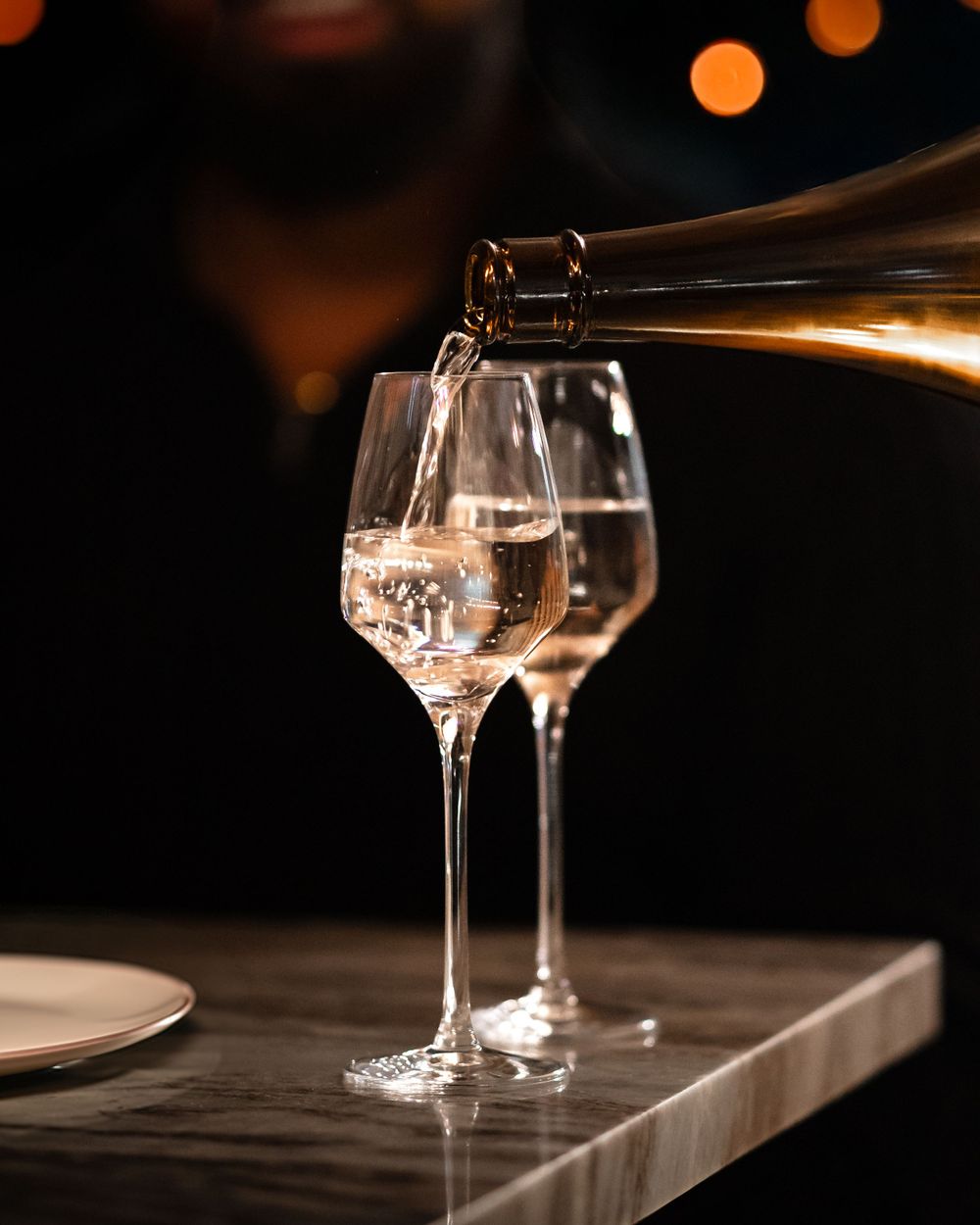 White wine being poured into elegant stemmed glasses against dark background with ambient lighting. Mtlshutter photography