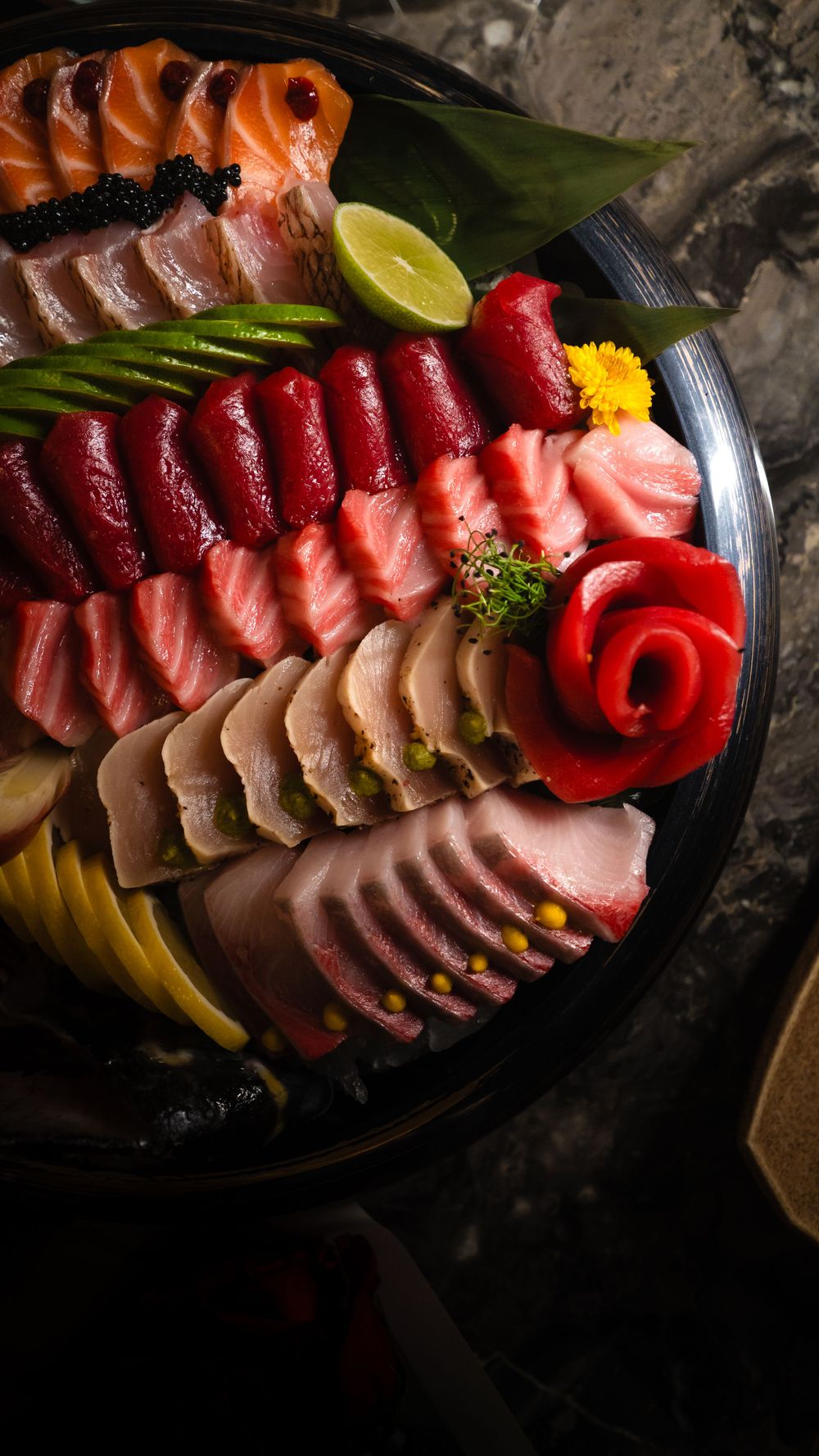 Colorful assortment of fresh sashimi slices arranged with garnishes and citrus on a dark plate. Mtlshutter photography