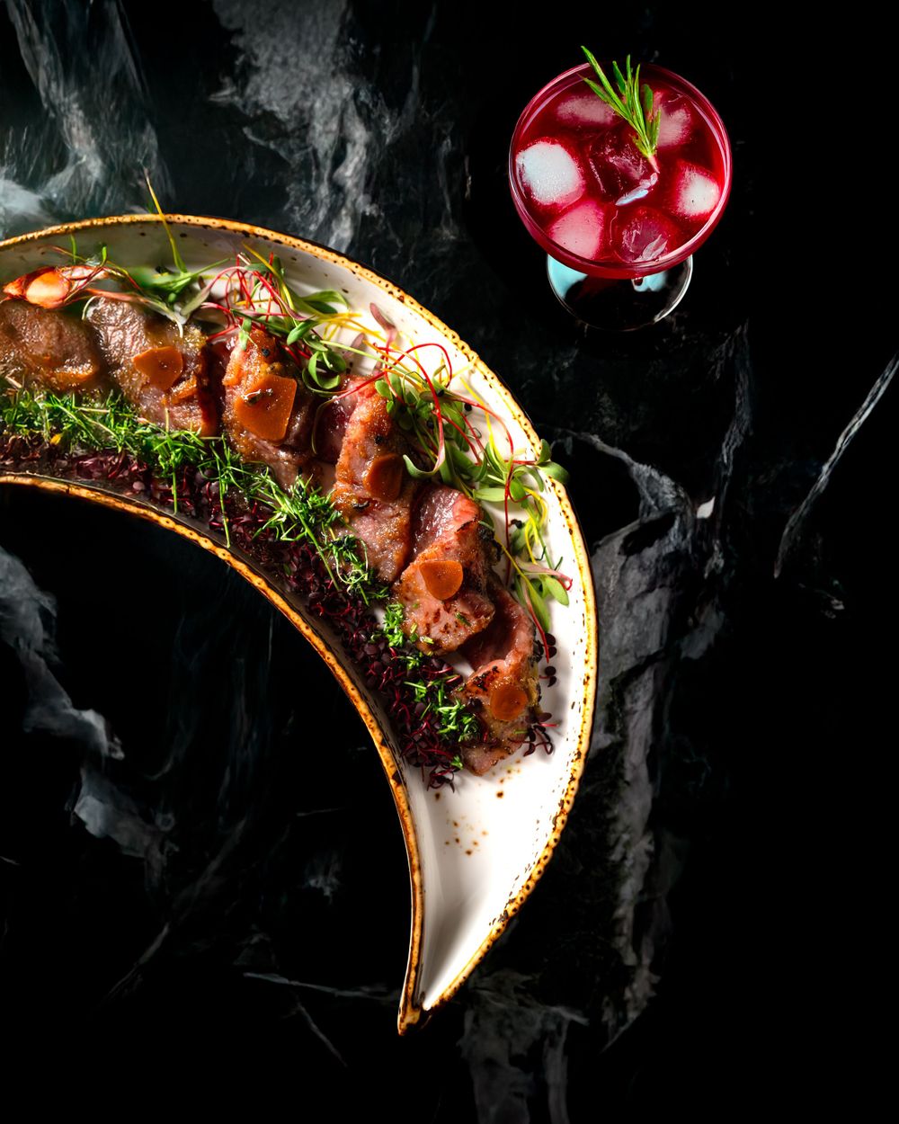 Artistically plated meat dish with microgreens on curved ceramic plate next to red cocktail. Mtlshutter photography