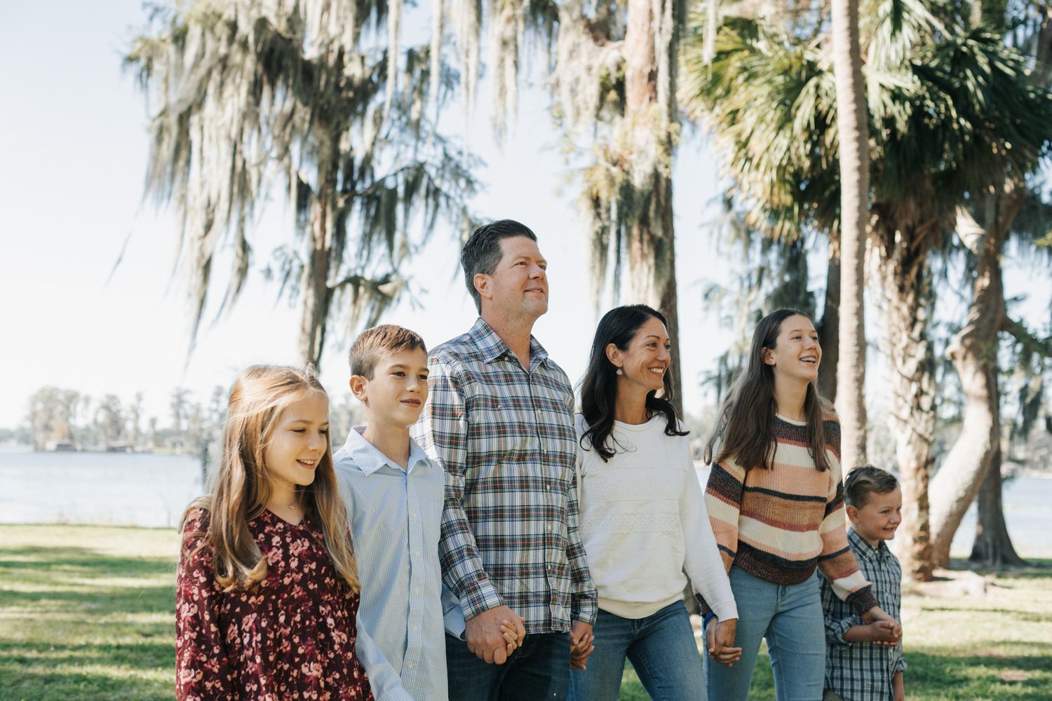 A family of six holds hands and walks together on a grassy lakeside lawn shaded by palm trees and Spanish moss.