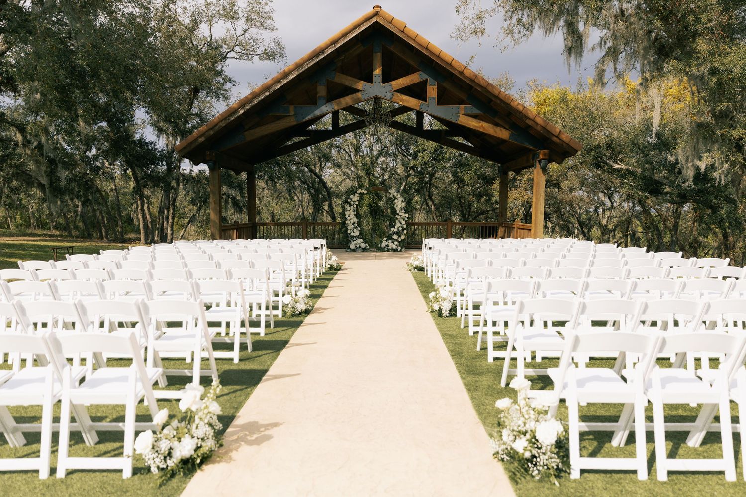 A wedding ceremony site with rows of white chairs along a grassy aisle leading to a rustic wooden pavilion beneath oak trees.