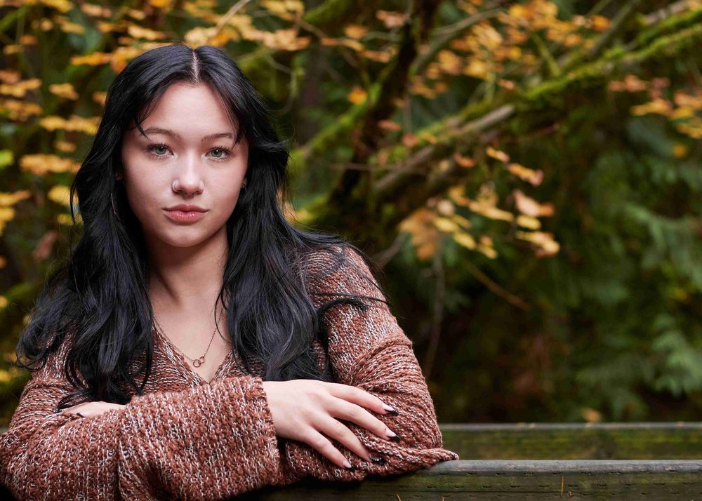 High school senior female wearing brown sweater but leaning with arms crossed on wooden fence at Wildwood Park in Puyallup, WA.