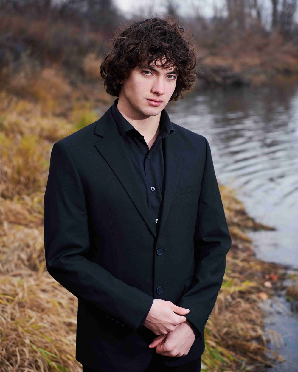 High school senior male in black suit, adjusting cuff while standing on bank near Okanogan river.