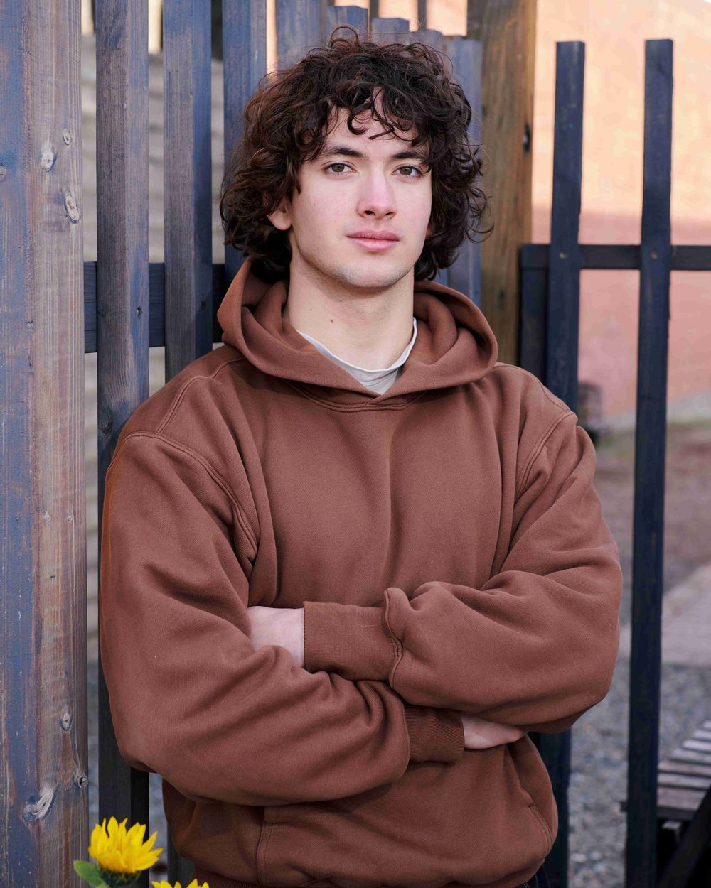 High school senior male wearing brown sweatshirt standing with arms crossed, leaning against brown wooden fence.