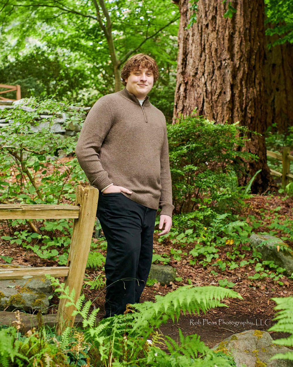 High School senior male wearing brown sweater while leaning against wooden fence on grounds at Lakewold Gardens in Lakewood, WA.