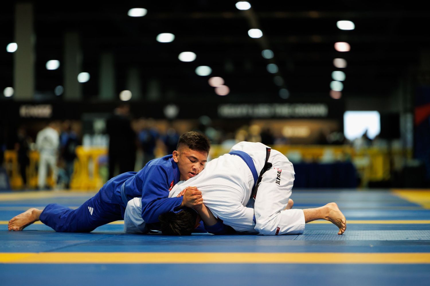 Competitors in blue and white gis grappling on dark mats during a tournament.
