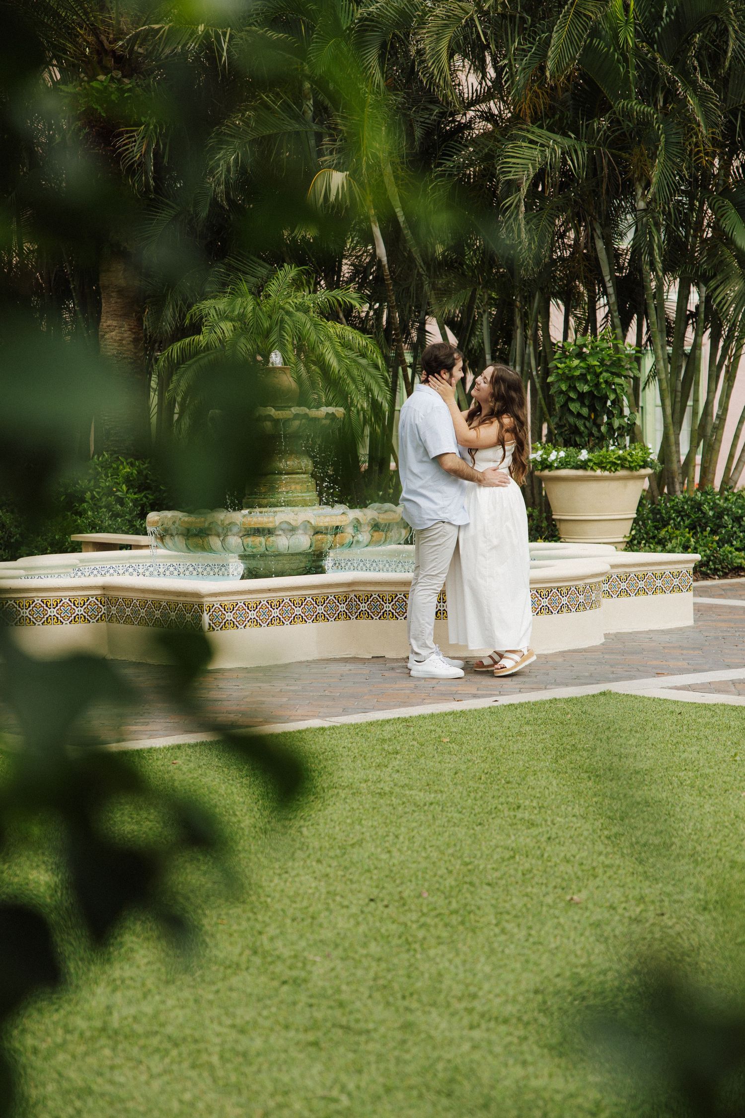 Couple in white outfits embracing by a fountain in a lush tropical garden setting.
