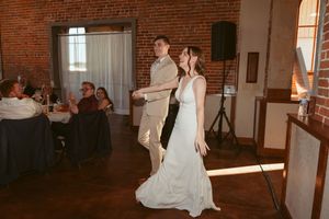 Bride in flowy white wedding dress twirls gracefully in a rustic brick-walled venue with warm lighting.