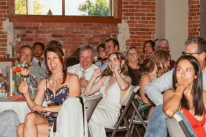 Wedding guests react emotionally while seated at round tables in a brick-walled reception venue.