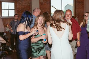 Party guests in formal attire including green and blue dresses dance and socialize at an event in a brick-walled venue.