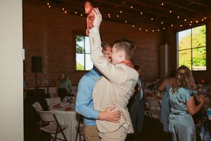 A joyful moment of dancing and lifting at a wedding reception in a rustic brick venue with string lights.