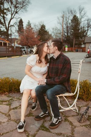 A romantic couple sits together on a white bench surrounded by fall foliage in a parking lot setting.