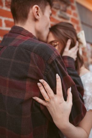 Close-up shot showing a hand with engagement ring resting on a burgundy plaid flannel shirt against brick wall.