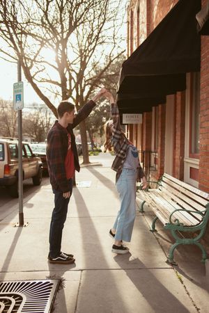 A couple dances playfully on a sunlit sidewalk in front of vintage storefronts with black awnings.