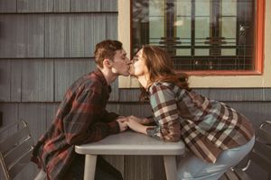 Two people in plaid shirts share a romantic moment across a table outside a building with gray siding.
