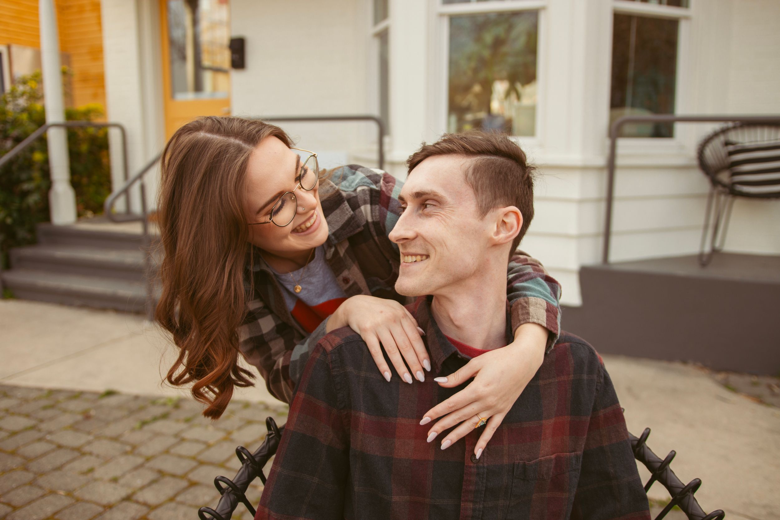 Happy couple embracing and laughing together outdoors with stone wall and building in background.