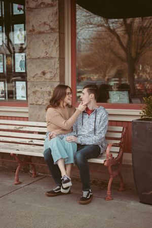 Playful couple sitting on wooden bench sharing affectionate moments against stone building.