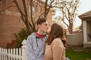 A couple shares a tender kiss in front of a white picket fence at sunset.