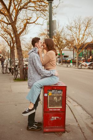 Romantic couple embracing on a red newspaper box on a street lined with autumn trees.