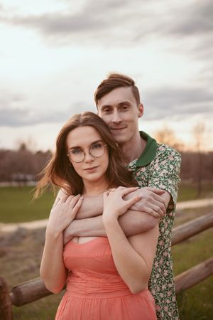 Couple in romantic pose at sunset with woman in pink dress and man in floral shirt against wooden fence.