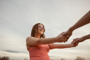 Woman in coral dress smiling while holding hands with someone against cloudy sky at sunset.