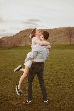 A couple embraces and spins on a green grassy hilltop at sunset, surrounded by rolling hills in the background.