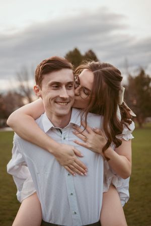 A romantic couple shares a playful piggyback ride moment in a grassy field at dusk.