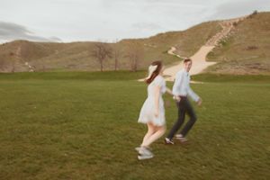 Two people in wedding attire run playfully through a green hillside field at sunset creating a romantic silhouette.