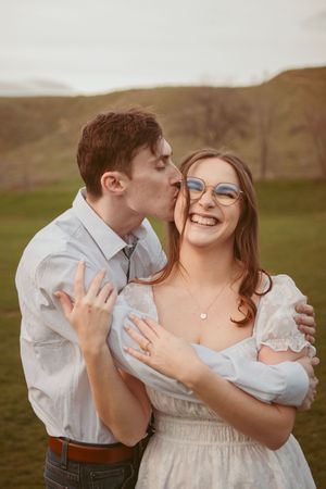 A couple in white embraces joyfully in a romantic outdoor engagement shoot at golden hour on rolling hills.