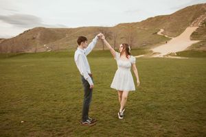 A couple dances in a white dress and formal attire on a grassy hillside with mountains in the background.