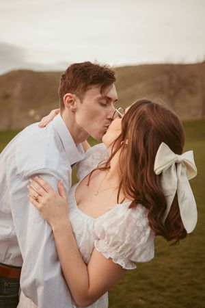 A romantic couple share a tender kiss in a white outfit and lace dress against rolling hills at sunset.
