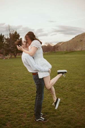 A couple in casual attire embraces and dances playfully in a grassy field during sunset.