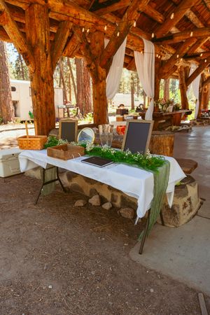Rustic outdoor dining area under wooden beams with white tablecloths and stone seating arrangements in a natural setting.