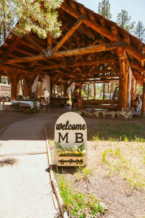 Welcome sign and rustic log pavilion entrance with M B lettering at a mountain resort venue.