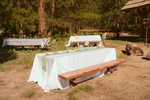 White banquet tables decorated with floral centerpieces and sage green runners set up outdoors in a natural wooded setting.