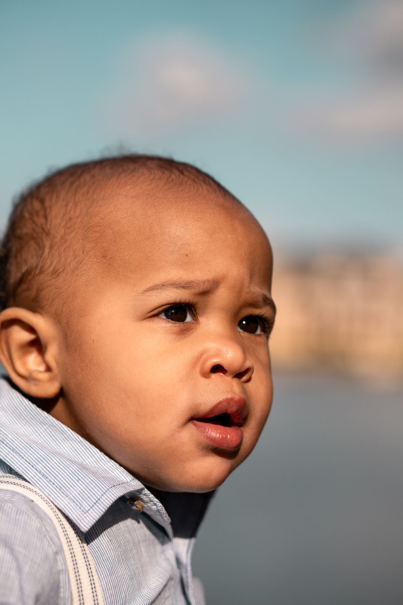 A young child wearing a light colored outfit looks to the side with an inquisitive expression against a blurry blue background.