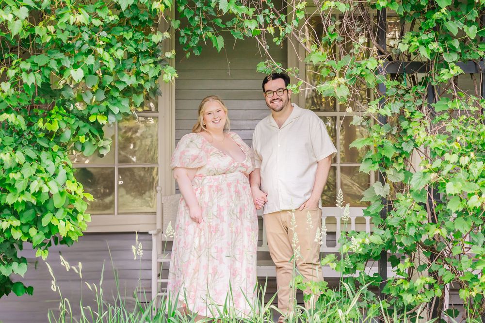 Couple stands together smiling on a lush garden patio with green vines growing on trellis in the background.