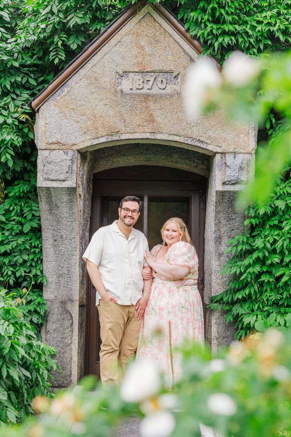 A couple poses together in front of a historic stone archway covered in lush green ivy in a garden setting.