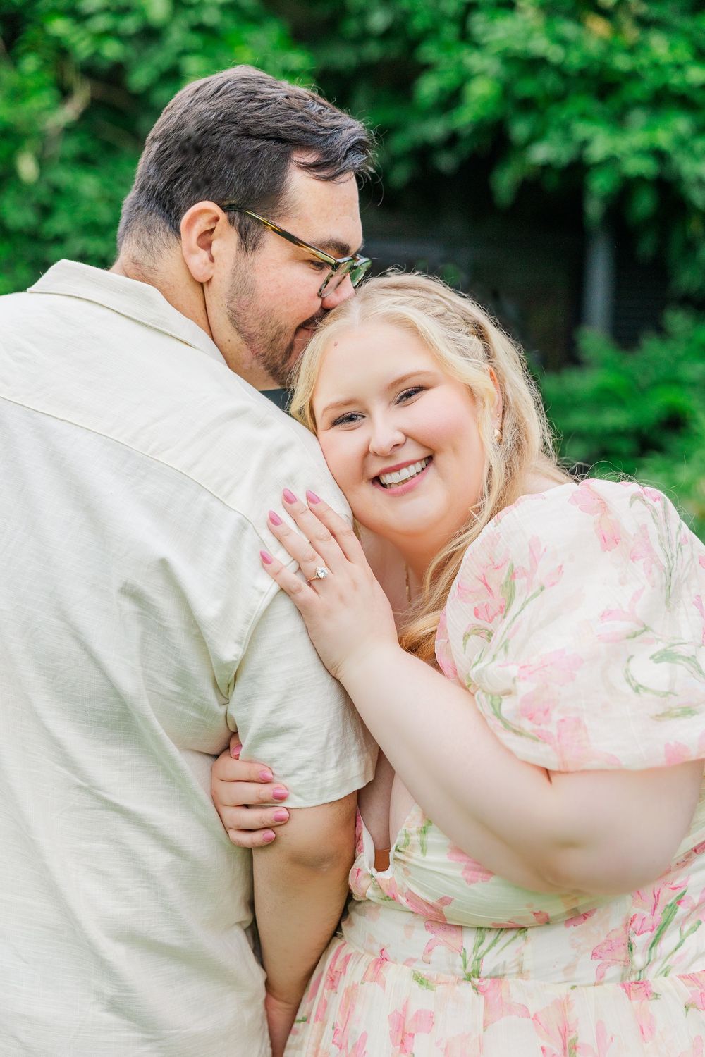 A romantic couple embraces outdoors, with her wearing a floral pink dress and him in a white shirt against green foliage.