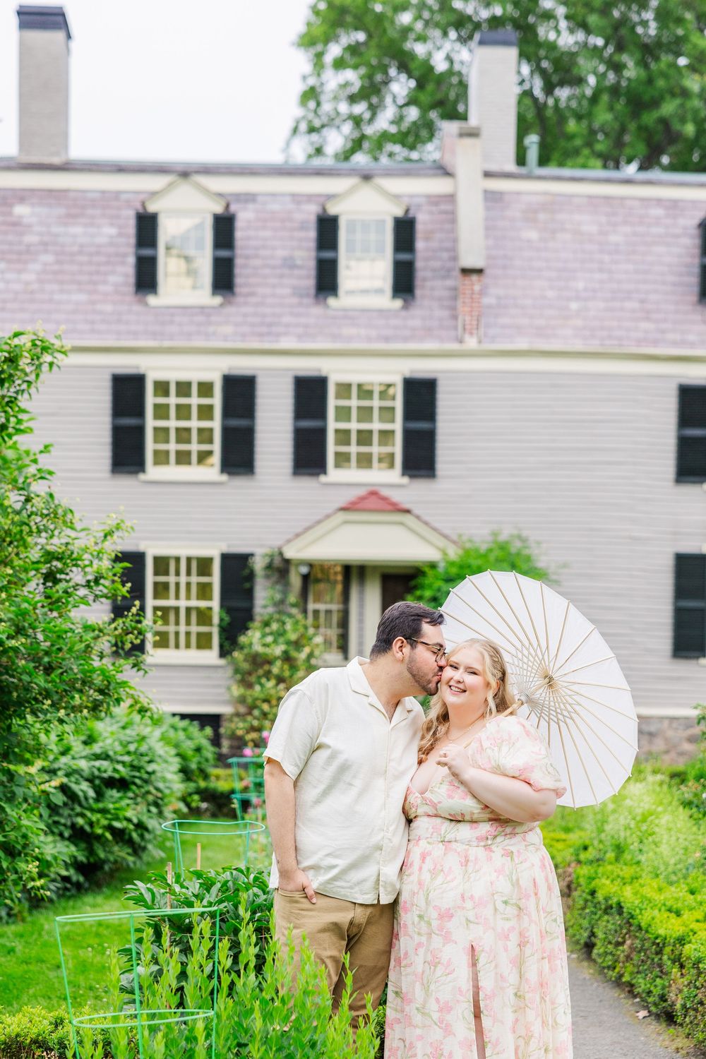 A couple shares an intimate moment with a white parasol in front of a classic colonial style house with black shutters.