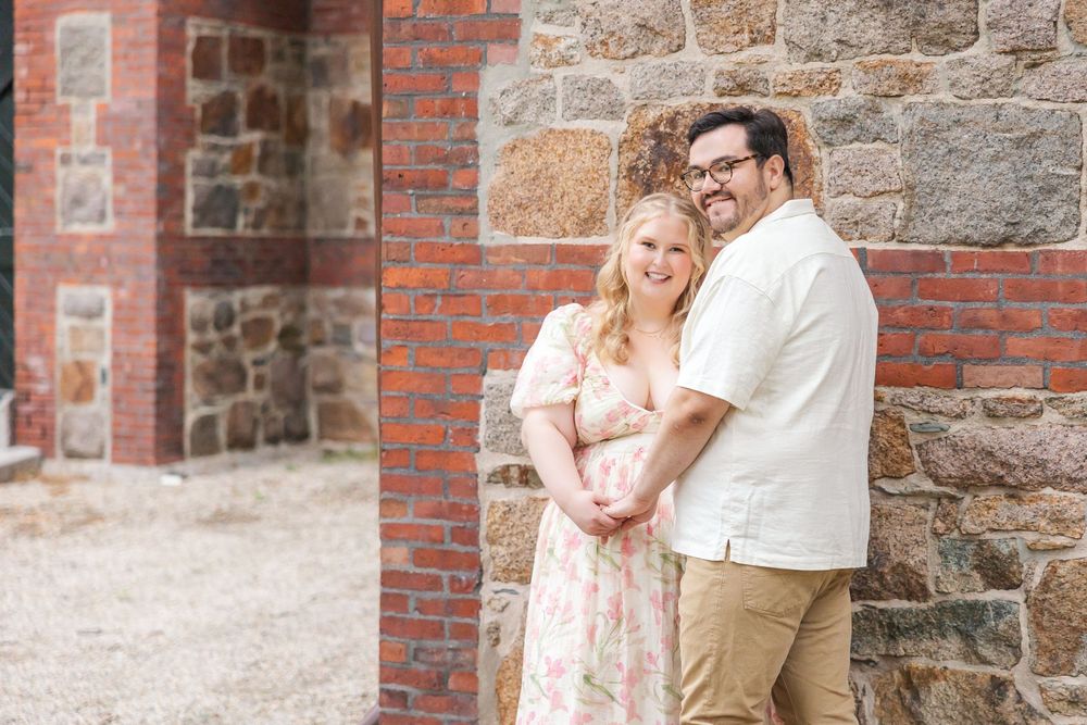 A couple embraces while standing next to a rustic stone and brick wall, the woman wearing a pink floral dress.